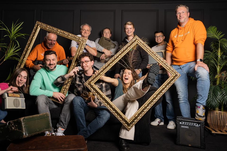 Het team van Younicorp poseert lachend met gouden lijsten, een gitaar en een oude telefoon in een fotostudio.