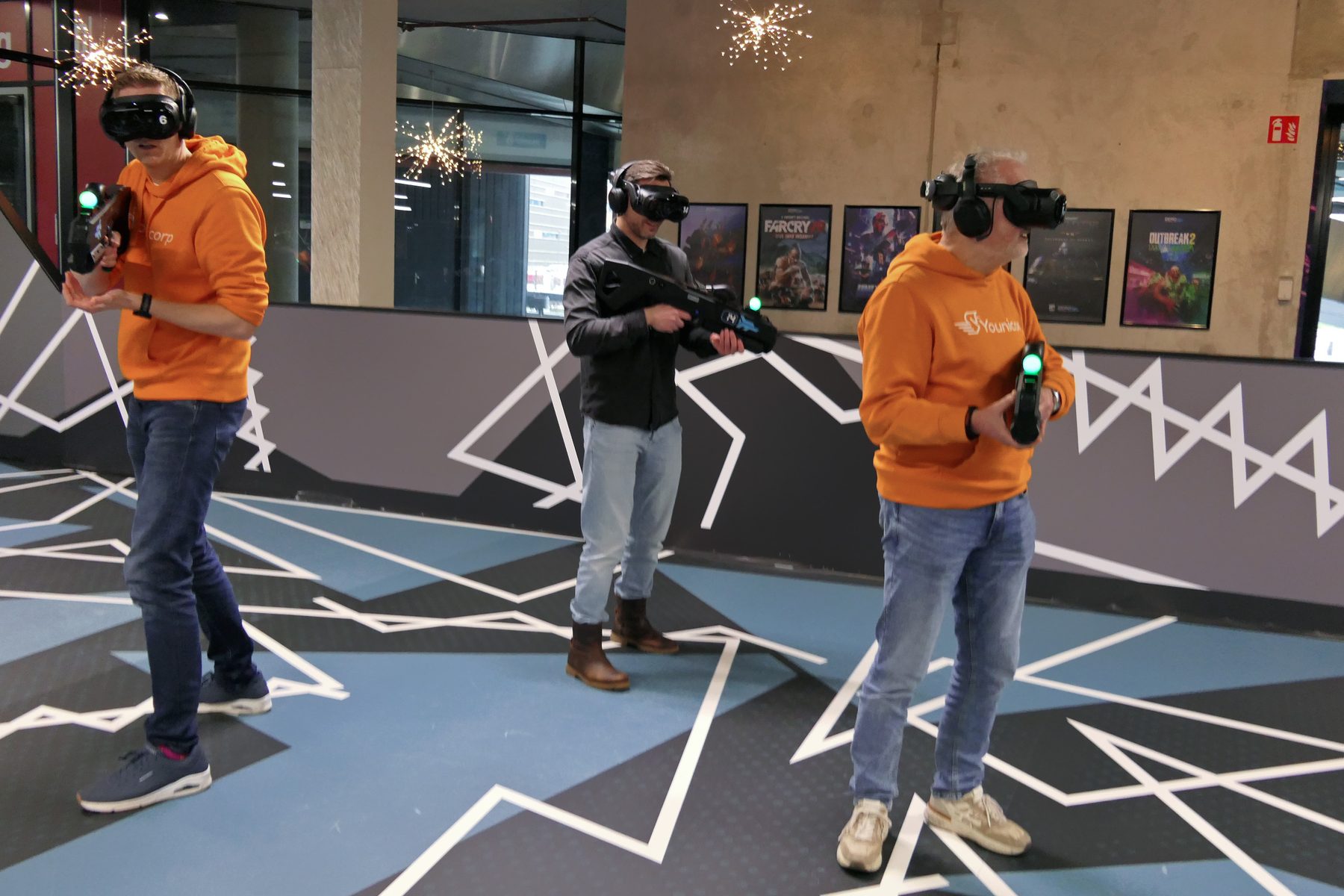Twee collega's in oranje hoodies met VR-brillen op een grafisch gevloerde arena.