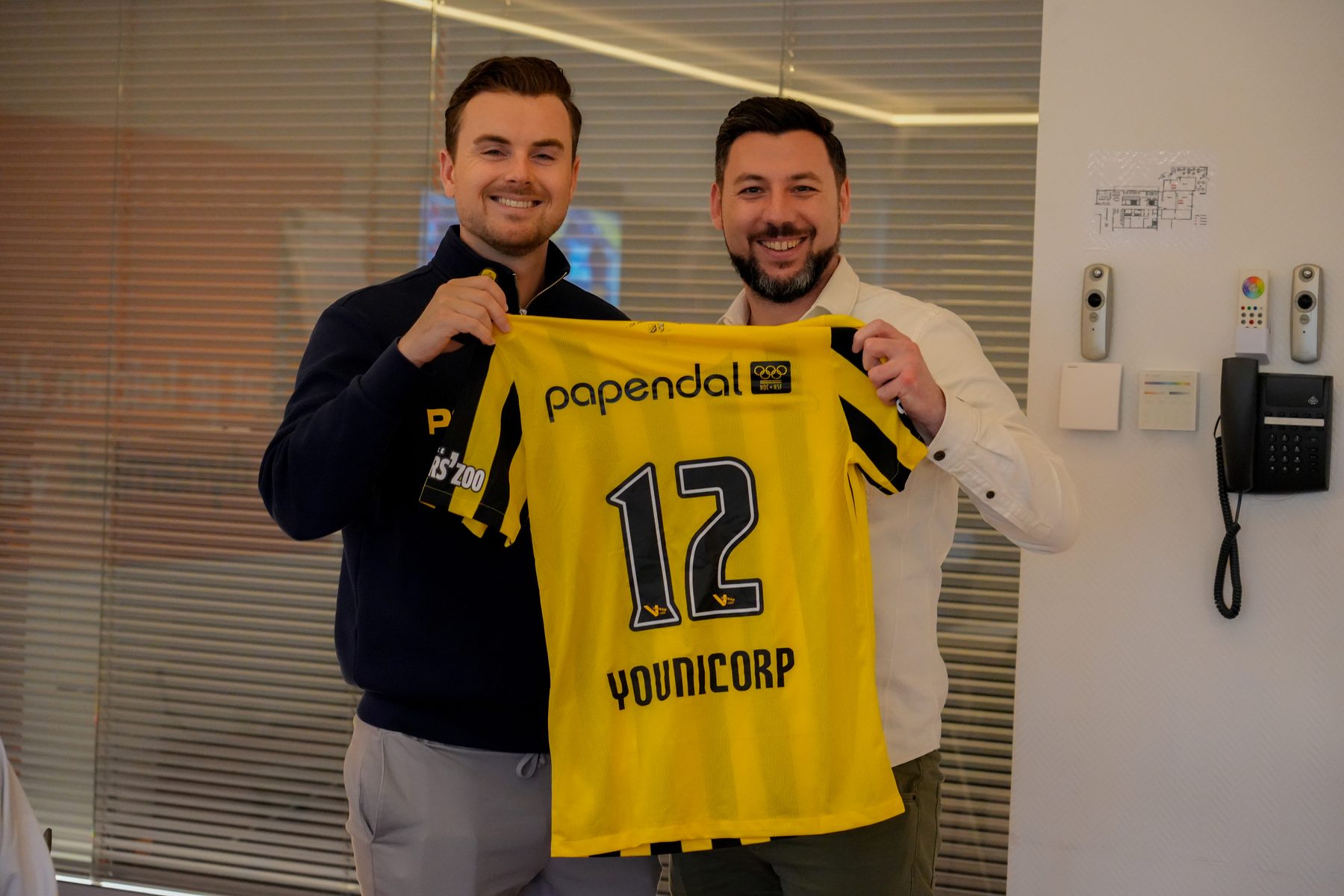 Twee mannen houden lachend een geel-zwart Vitesse-shirt omhoog met nummer 12 en Younicorp op de voorkant.