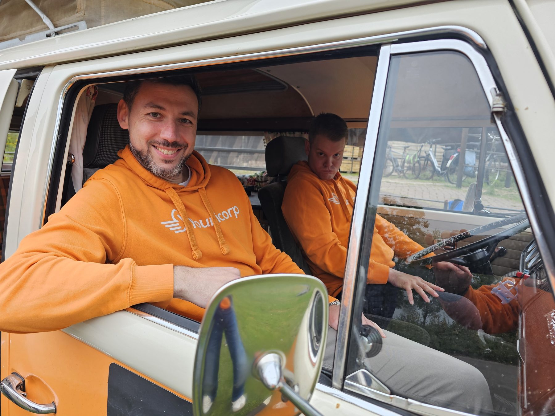 Een collega in een oranje Younicorp-hoodie leunt lachend uit het raam van een oud Volkswagen-busje.