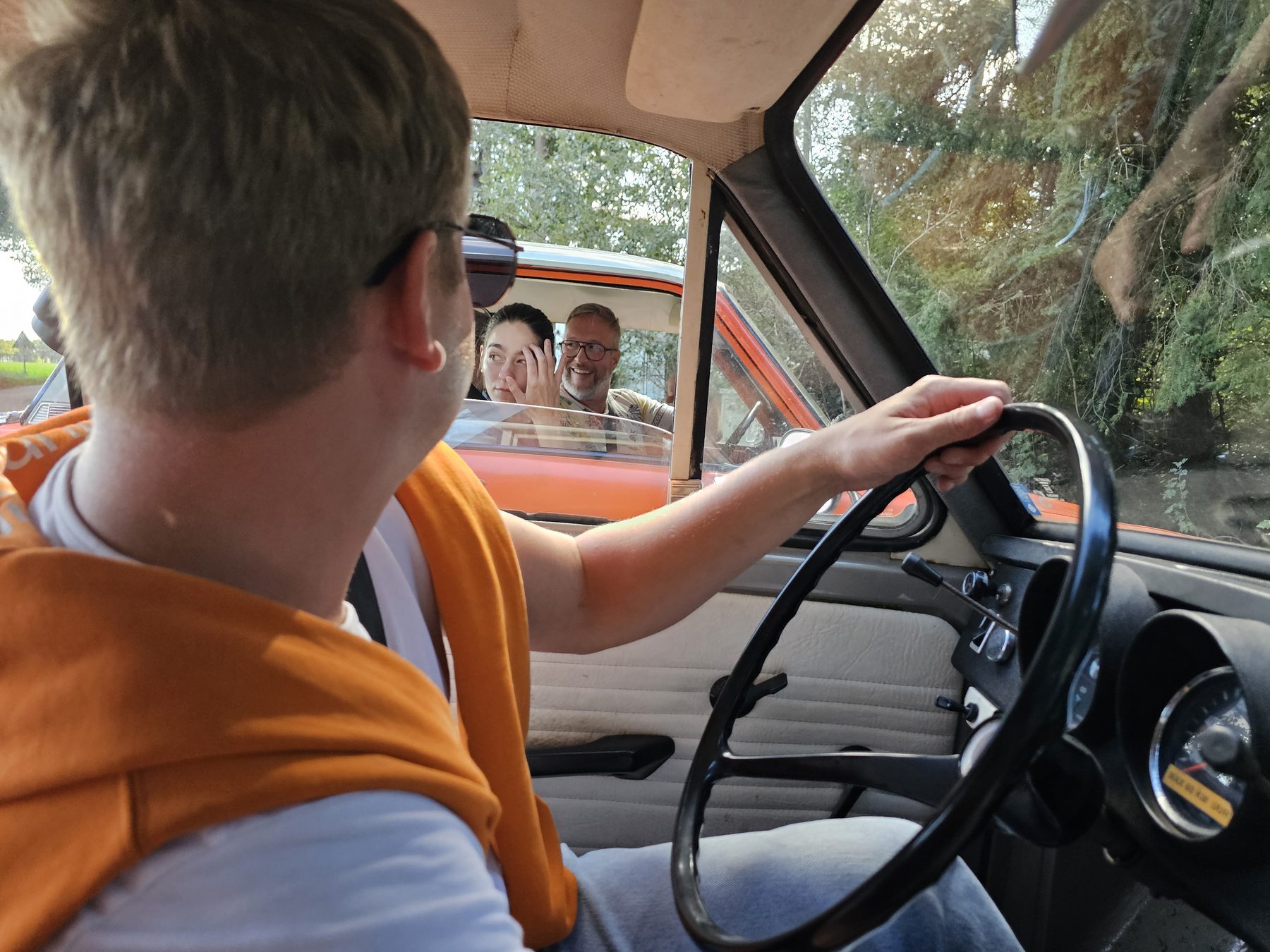 Vanuit een Dafje kijkt de bestuurder om naar collega's in de rode Daf ernaast, die lachend terugkijken.
