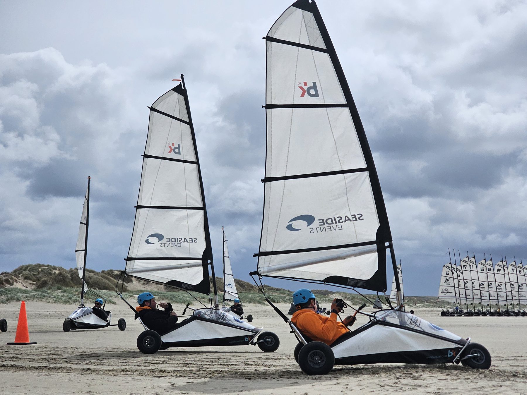 Strandzeilers met witte zeilen rijden over het strand onder een bewolkte lucht.