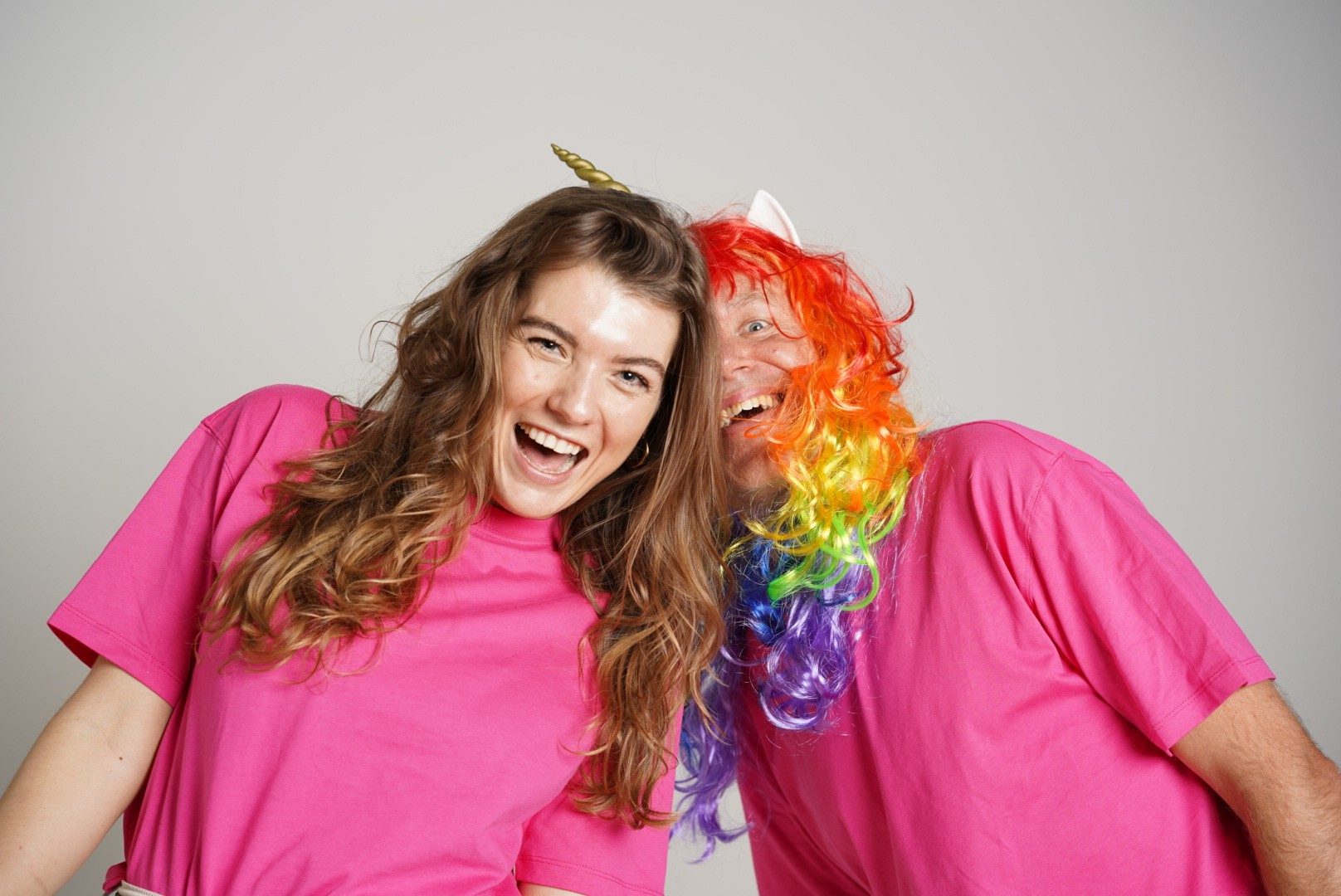 Twee collega's in roze shirts lachen hoofd tegen hoofd; een van hen draagt een regenboogpruik met eenhoorn.