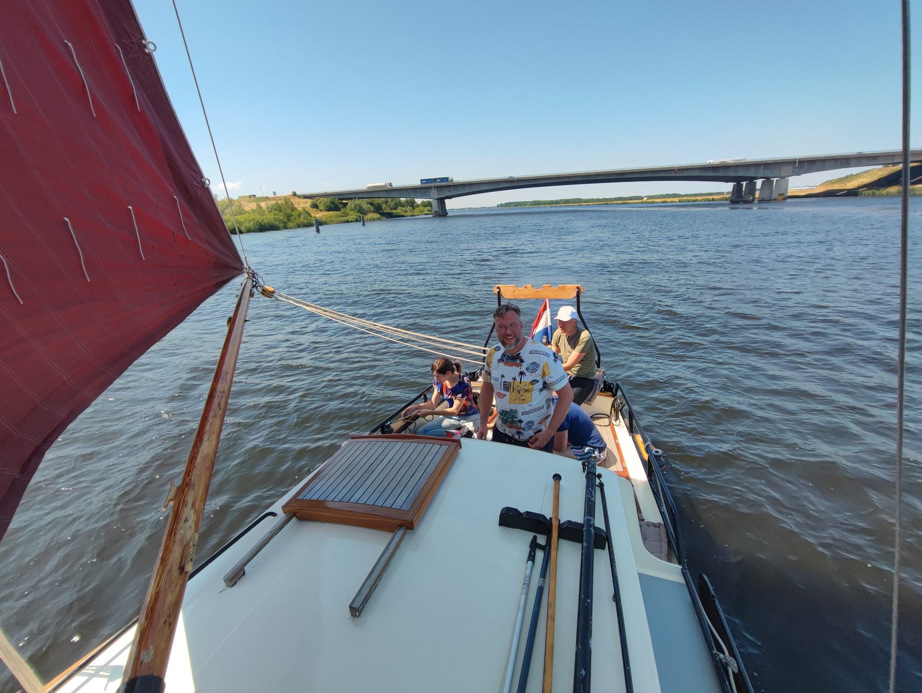 Vanaf een zeilboot met rood zeil kijk je over de rivier naar een brug.