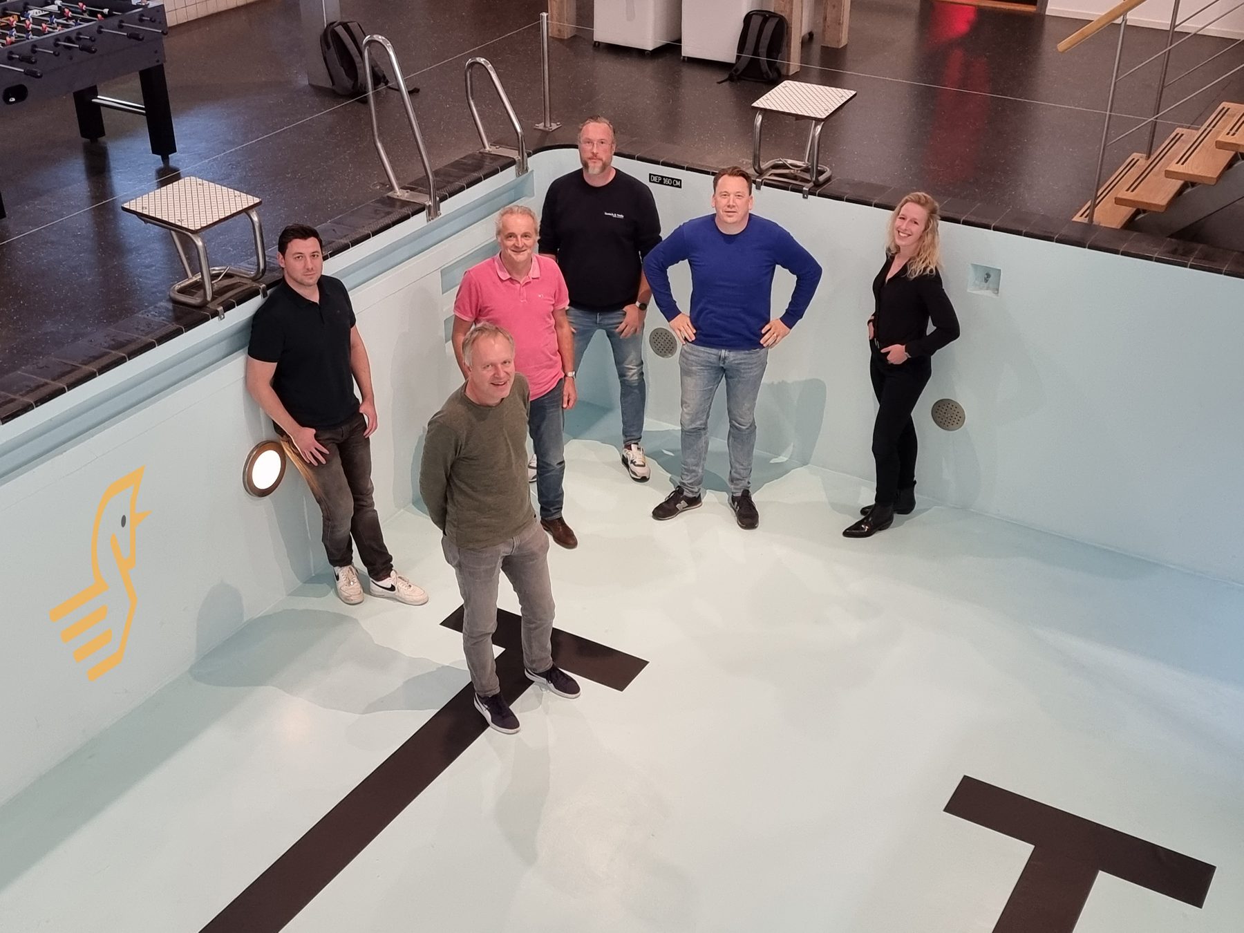 Het team staat in een leeg, drooggelegd zwembad met witte tegels.