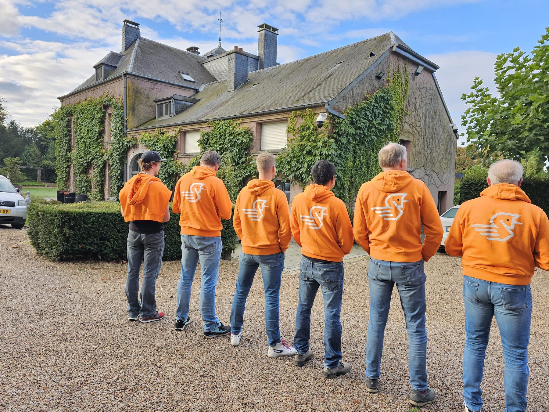 Zeven collega's in oranje Younicorp-hoodies staan met de rug naar de camera voor een oud landhuis.