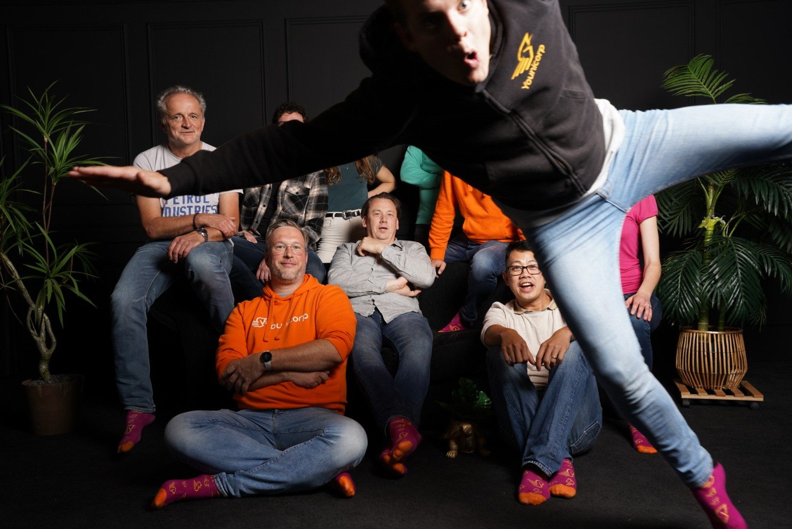Groepsfoto in de studio op een zwarte achtergrond; een collega springt horizontaal door het beeld terwijl de rest verbaasd kijkt.