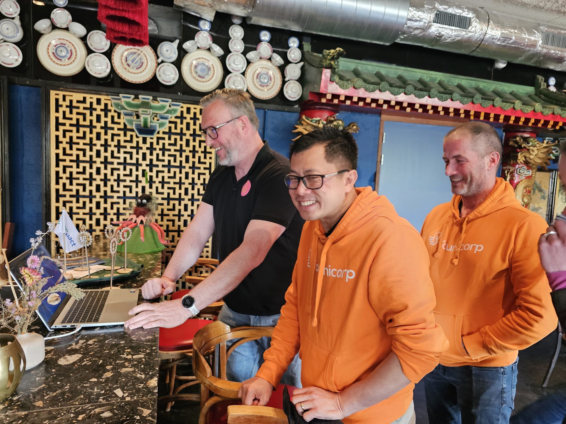 Drie collega's in oranje hoodies buigen zich samen over een laptop in een kleurrijk café.