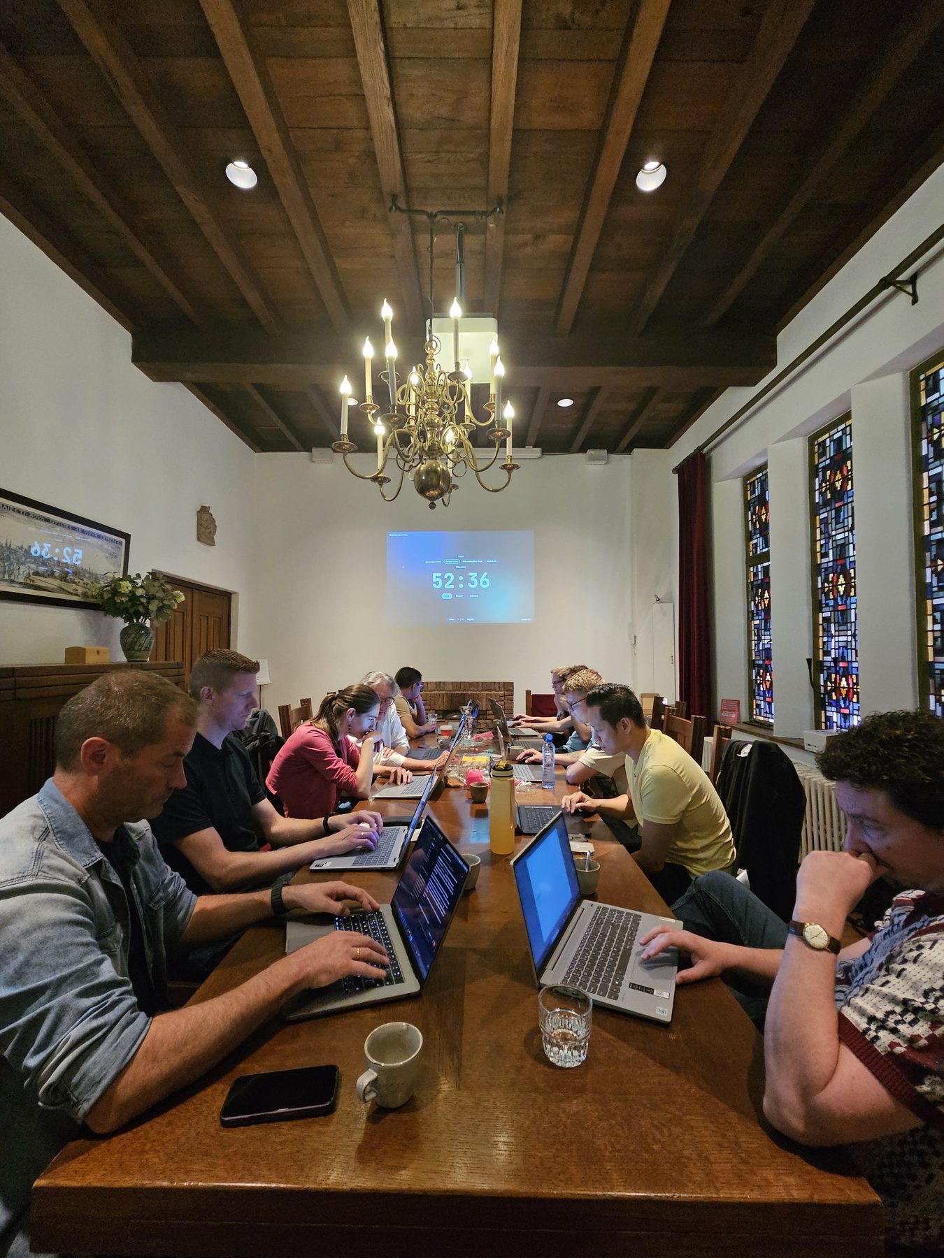 Collega's werken op laptops aan een lange tafel in een historische zaal met glas-in-loodramen.