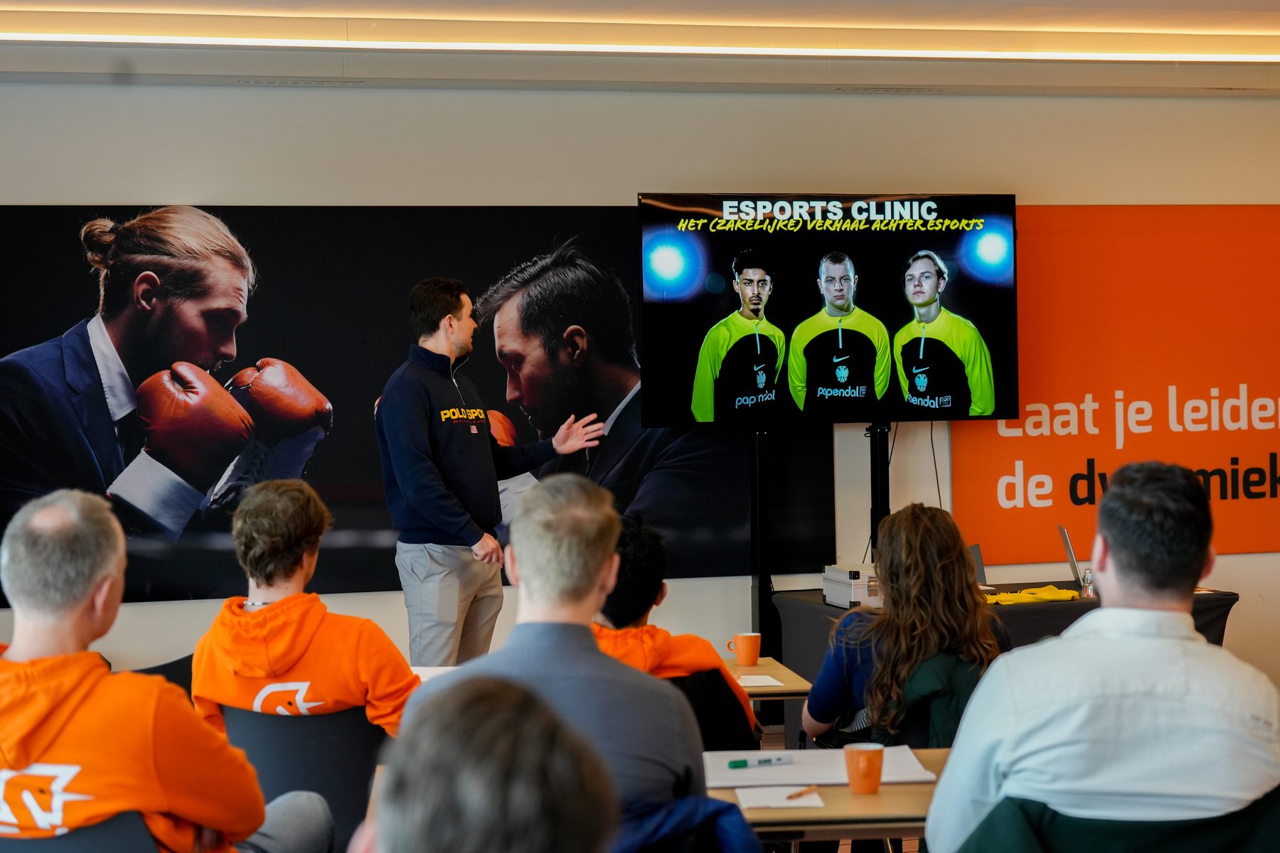 Een spreker staat voor de groep; op het scherm staat Esports Clinic met drie gamers in groene shirts.