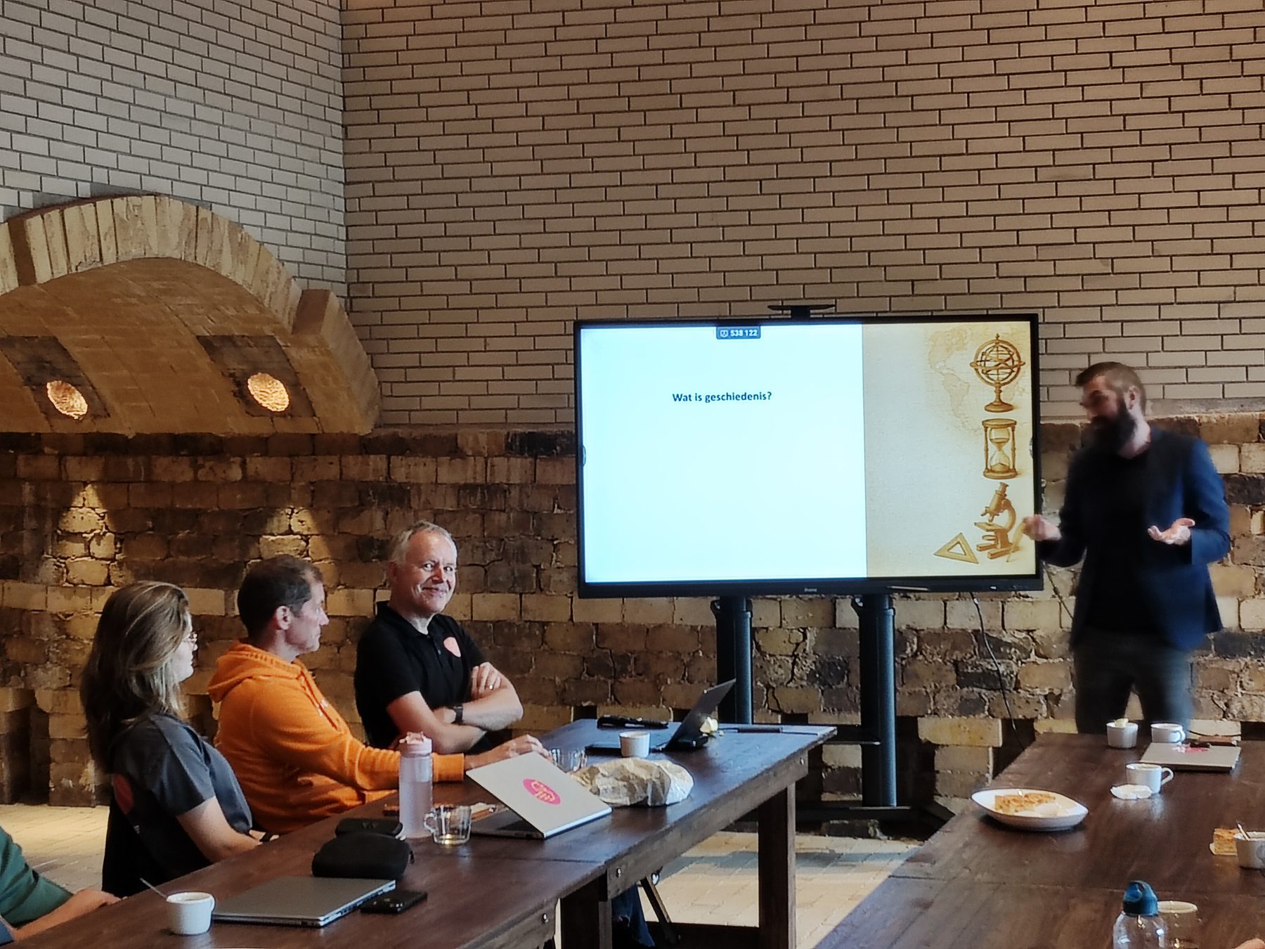 Een werksessie in een oud bakstenen gewelf, met een scherm en lange houten tafels.