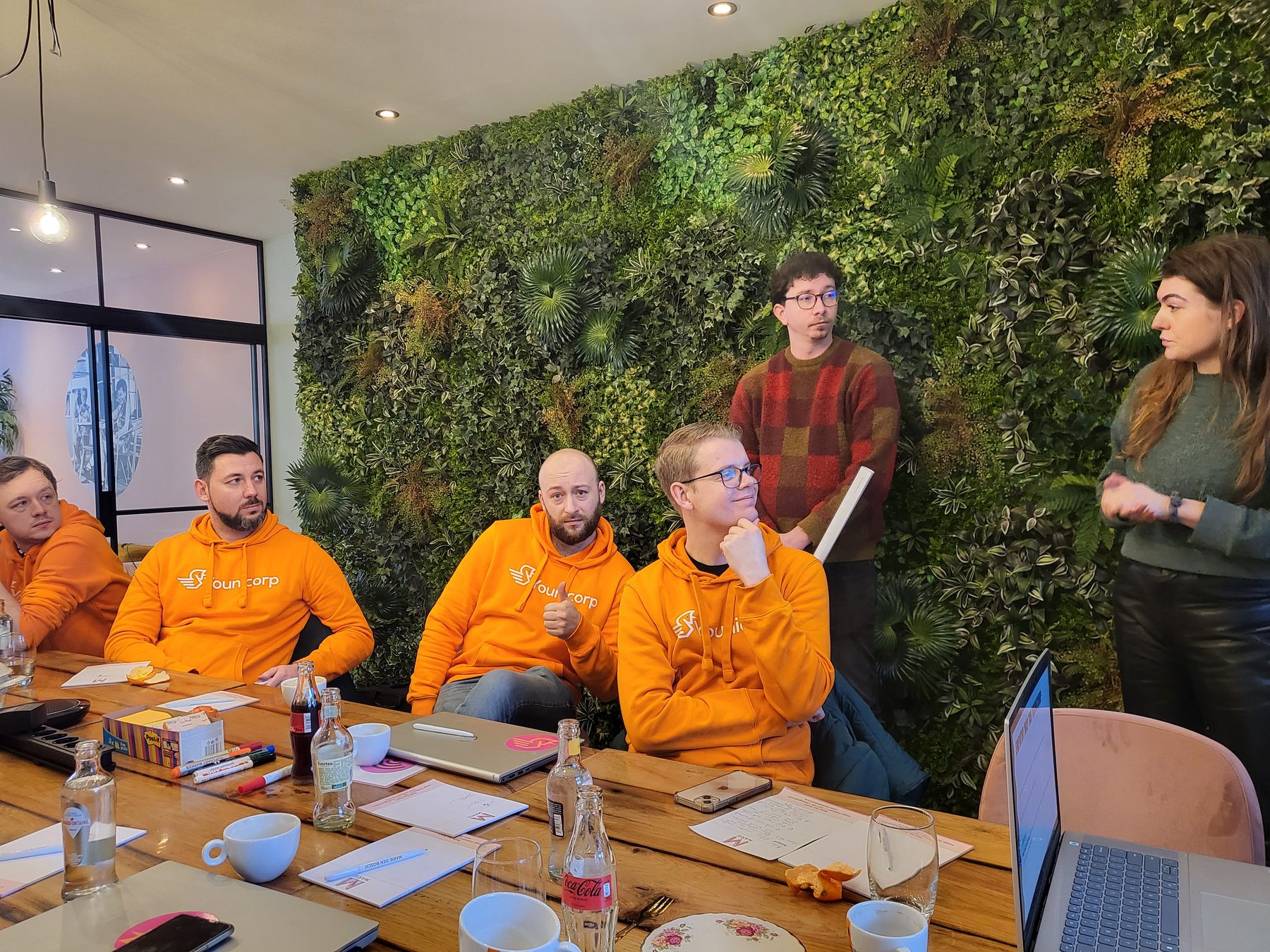 Collega's in oranje hoodies overleggen aan een tafel voor een groene plantenwand.