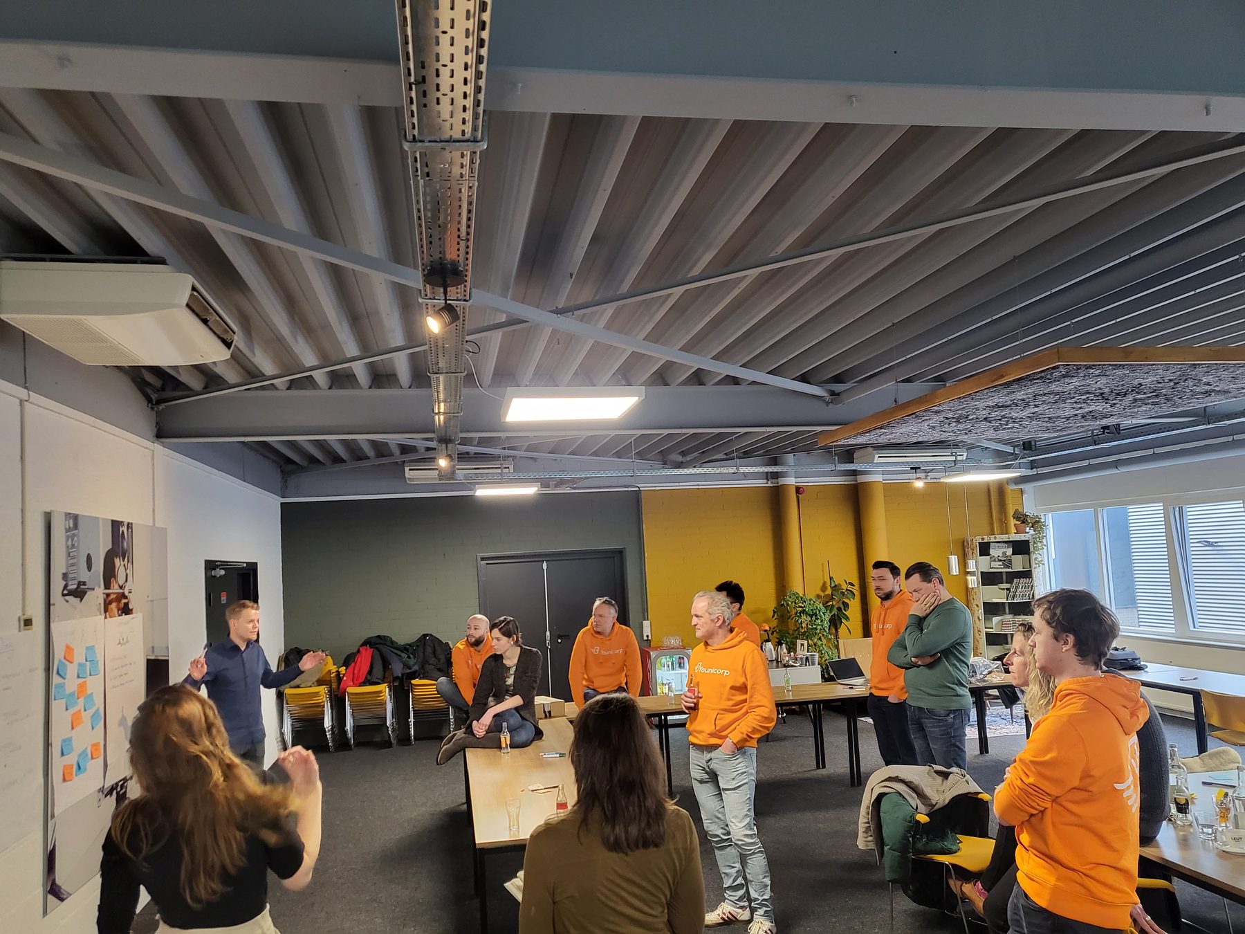 Een werksessie in een lichte zaal: collega's in oranje aan lange tafels, iemand presenteert.