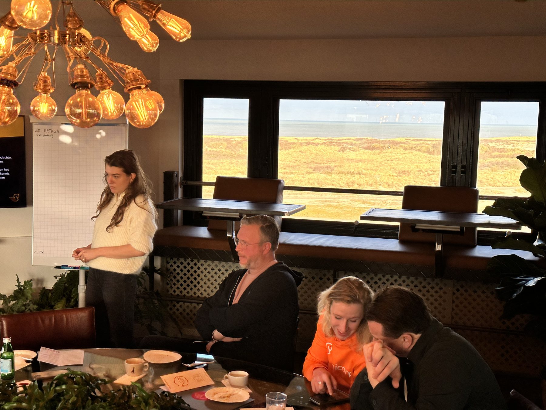 Een collega geeft een sessie bij een flip-over; het team luistert aan tafel, met uitzicht op de duinen.