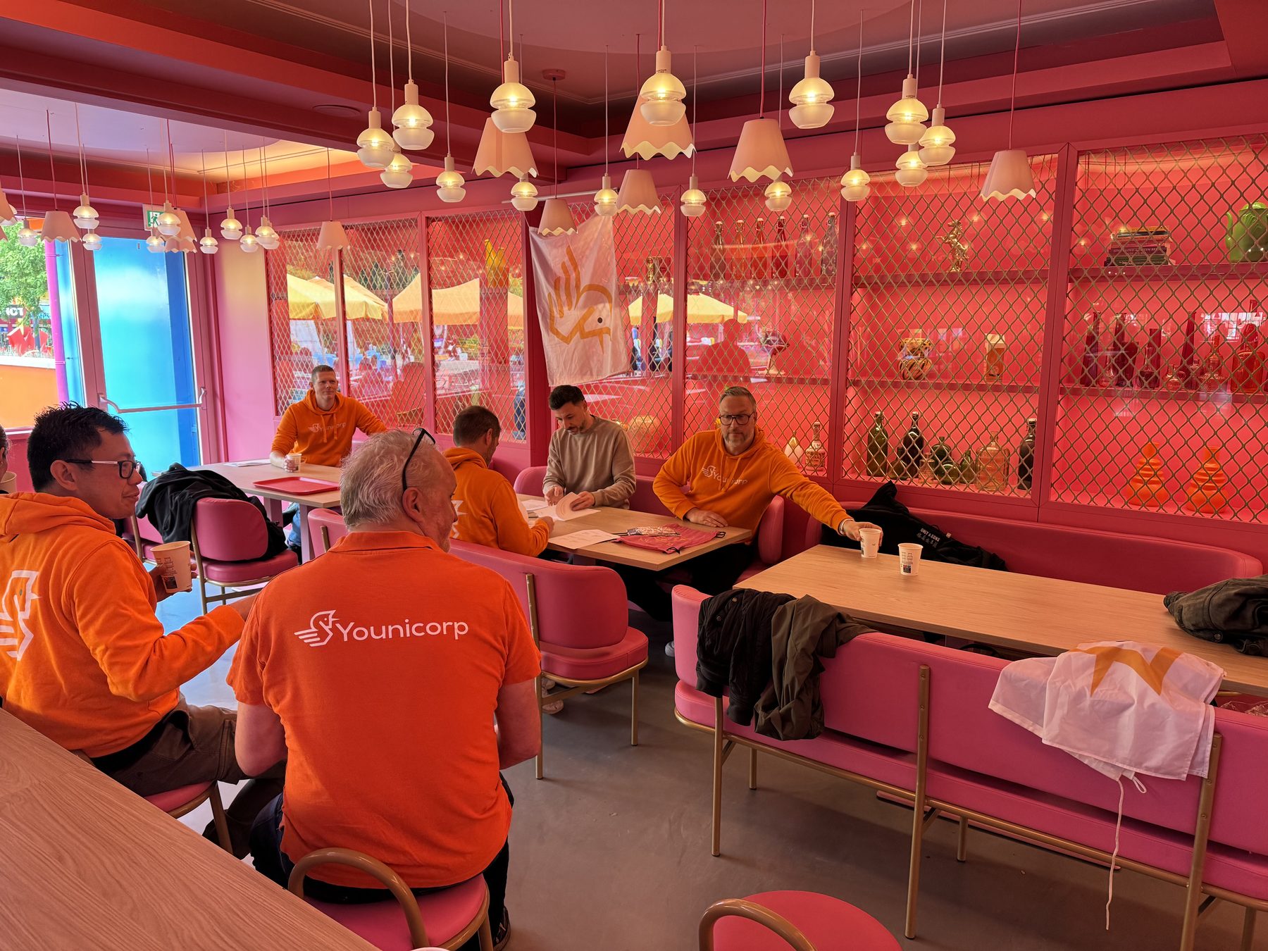 Collega's in oranje hoodies werken aan tafels in een roze ingericht restaurant.